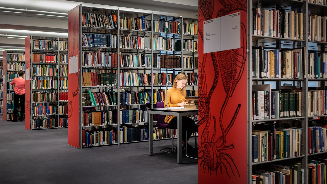 Photograph of a library with rows of free standing books shelves, filled with books. On the end of each row is a large red graphic plant like design and book category information signs. Between the first two rows a young woman is sitting at a table writing in a notebook, spotlit by a small desk lamp. In the distance another person can be seen standing at a bookshelf, back to camera.
