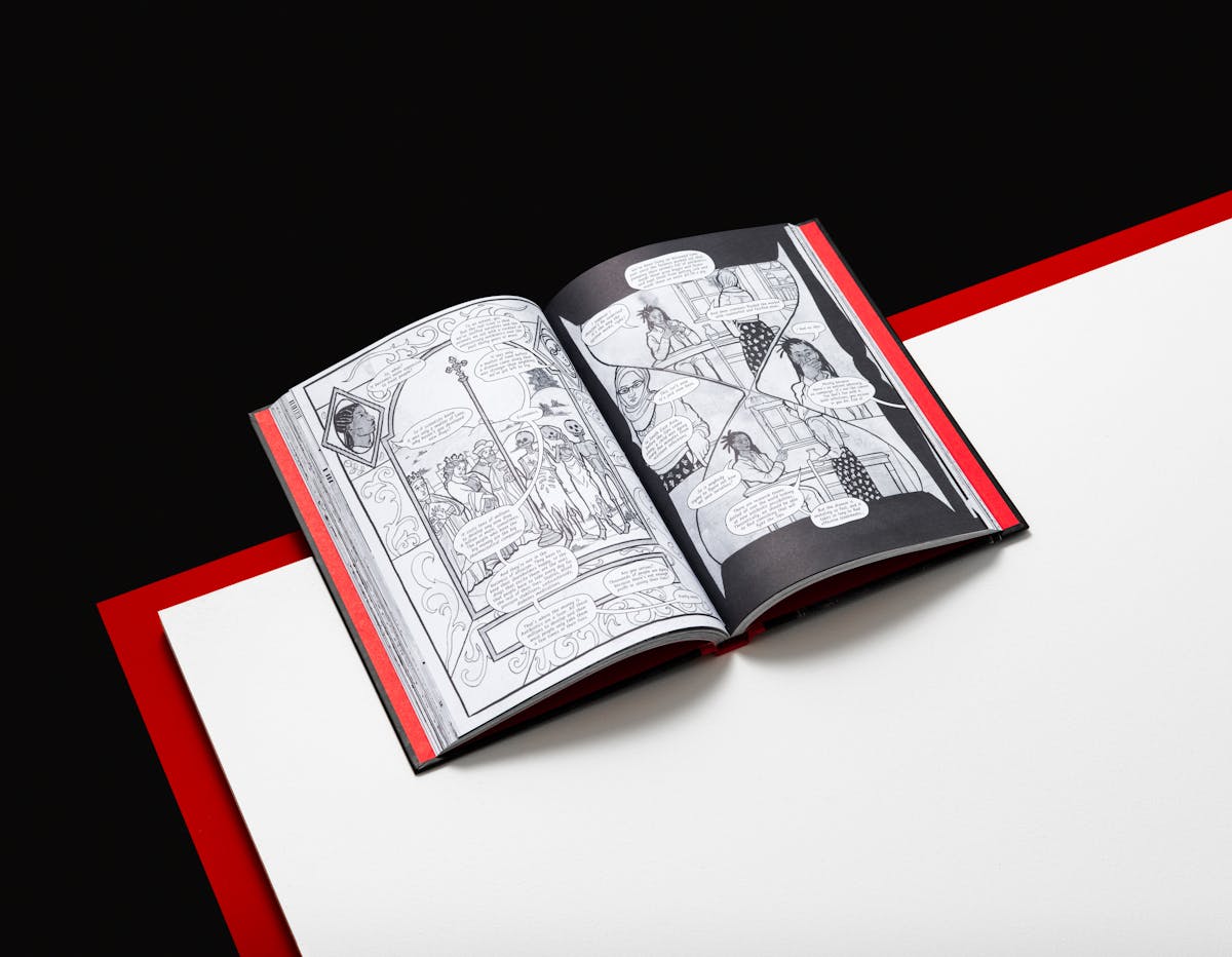 Photograph of an open hardback book resting on a white surface. The top part of the book is overhanging the white surface. Just below the white surface is another red surface creating an edge around the white top. This red compliments the red colour of the inside of the hardback cover. The two open pages of the book show black and white graphic novel illustrations. Behind the book and the surfaces is a black background.