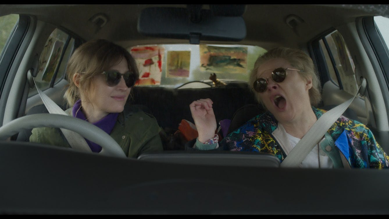 Film still from 'Typist Artist Pirate King' showing a view from a car dashboard of two women driving in a car wearing sunglasses. 