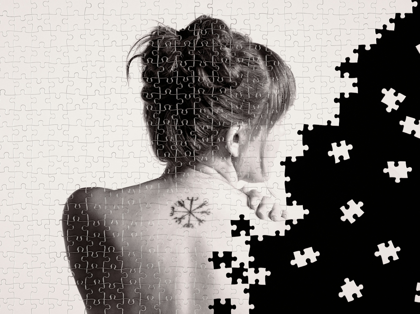 Photograph of a part made jigsaw puzzle containing hundreds of pieces. The jigsaw has been photographed against a black background, with many of the unused pieces scattered around in the black space. The image on the jigsaw is a warmed toned, monotone photograph of a nude woman against a plain light background. The woman is pictured from behind, showing the top half of her back and her head which is turned to the right. She has long hair tied in a bun and her left hand can been seen resting on her right shoulder. Between her shoulder blades is a tattoo in the shape of 4 long lines crossing each other at the centre of each line, with fork-like patterns on the end of each line. The missing jigsaw pieces create a black void which encroaches from the right side of the image, fragmenting the picture of the woman.