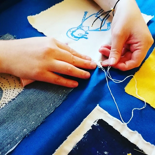A pair of hands sewing paintings on material squares onto a blue backing cloth.