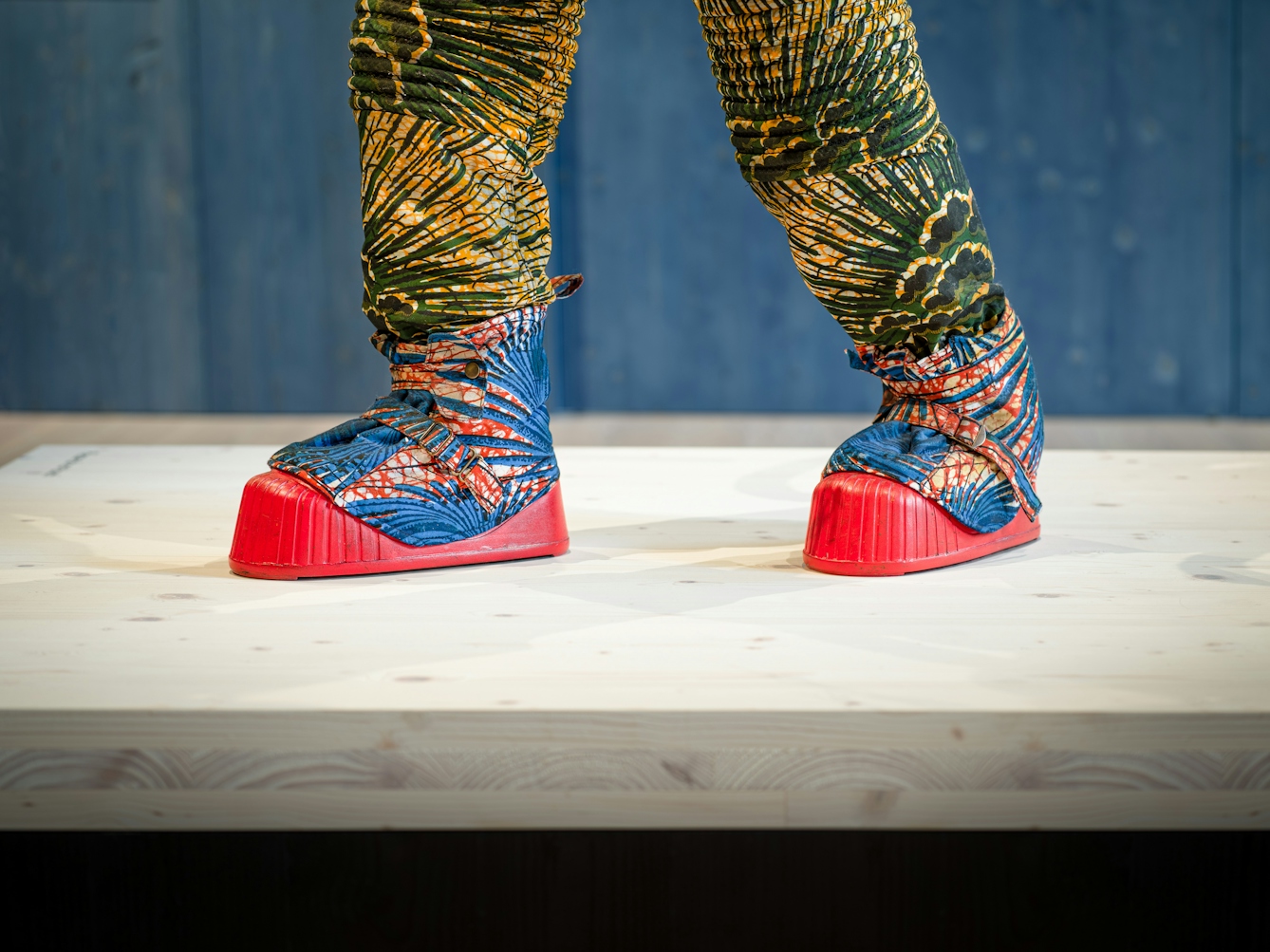 Photograph of an exhibition gallery space, with a blue stained wood wall in the background. In the foreground are the feet of a life-size artwork of a figure resembling an astronaut. The astronaut's boots are bright red in colour and their trousers are patterned in green and yellow.