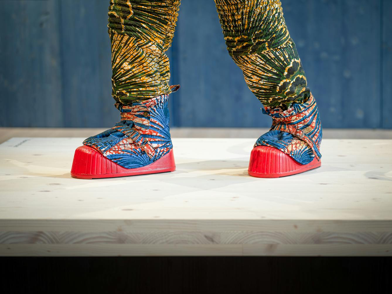 Photograph of an exhibition gallery space, with a blue stained wood wall in the background. In the foreground are the feet of a life-size artwork of a figure resembling an astronaut. The astronaut's boots are bright red in colour and their trousers are patterned in green and yellow.