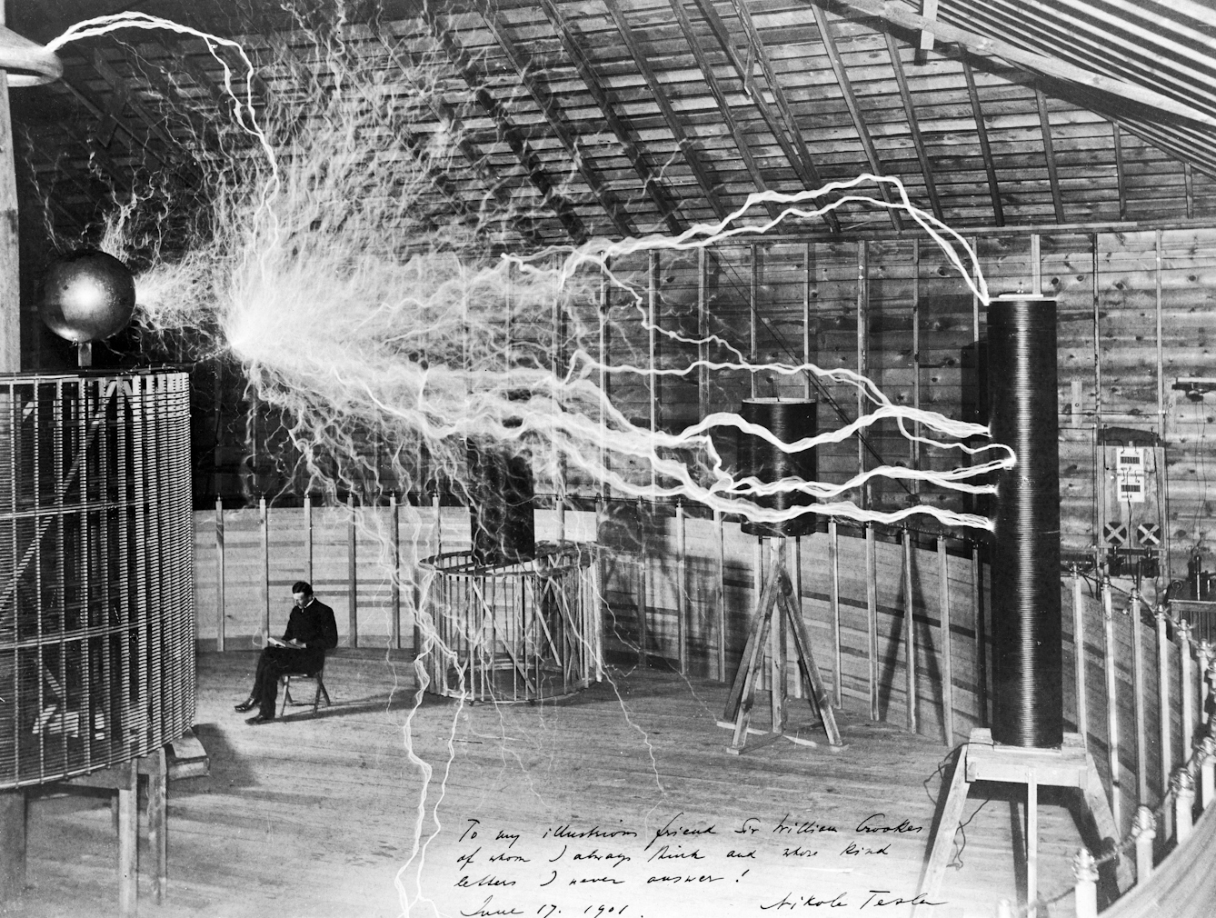 Black and white photograph of electricity arcing across a room with Nikola Tesla sitting in the background