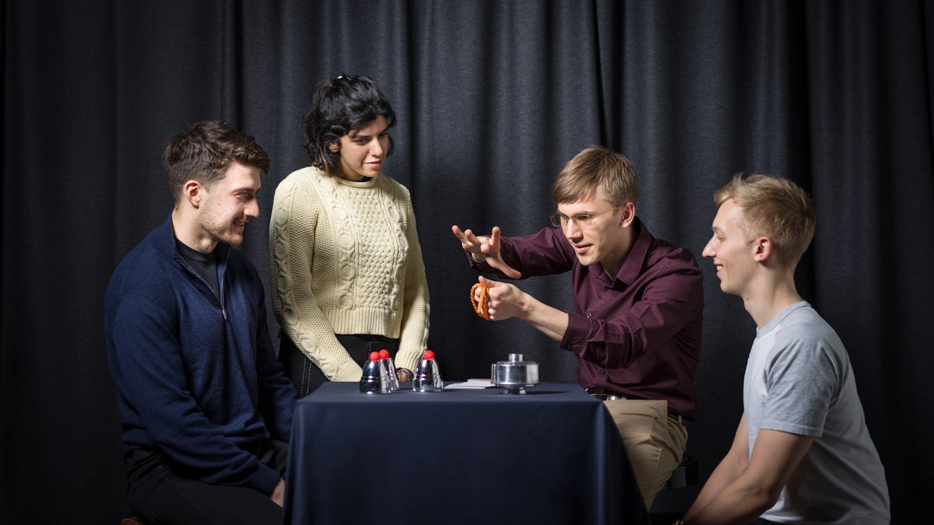 Photograph of a young woman and two young men gathered around a table, working with a magician to learn magic tricks. behind them is a grey curtain and on the table are magic props, such as cup and balls and a deck of playing cards.
