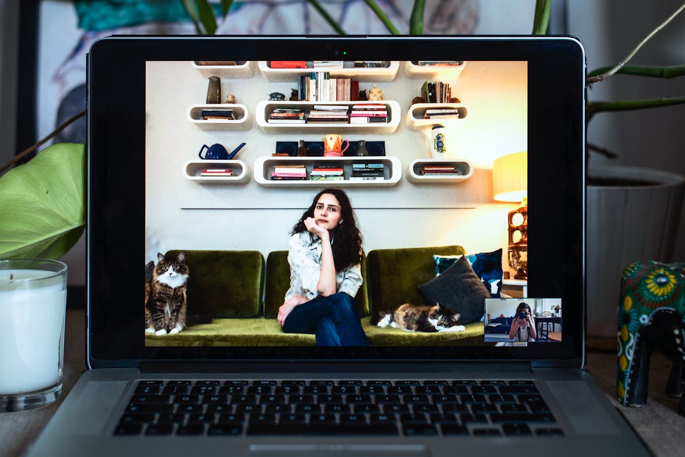 Photograph of an open laptop within a domestic scene. Most of the image is taken up with the screen, with part of the keyboard and trackpad visible. On the screen is a video call showing a woman sitting on her sofa with a cat either side of her. In the bottom right corner of the screen the photographer can be seen in a small  floating window, camera to her eye, in the process of taking a picture.