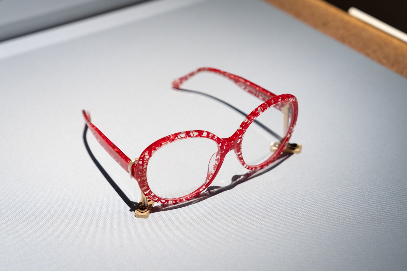 Photograph of a pair of red rimmed spectacles with the arms open, secured to a grey fabric display case base. The rims are transparent but covered in a web of red lines which criss-cross the frames.