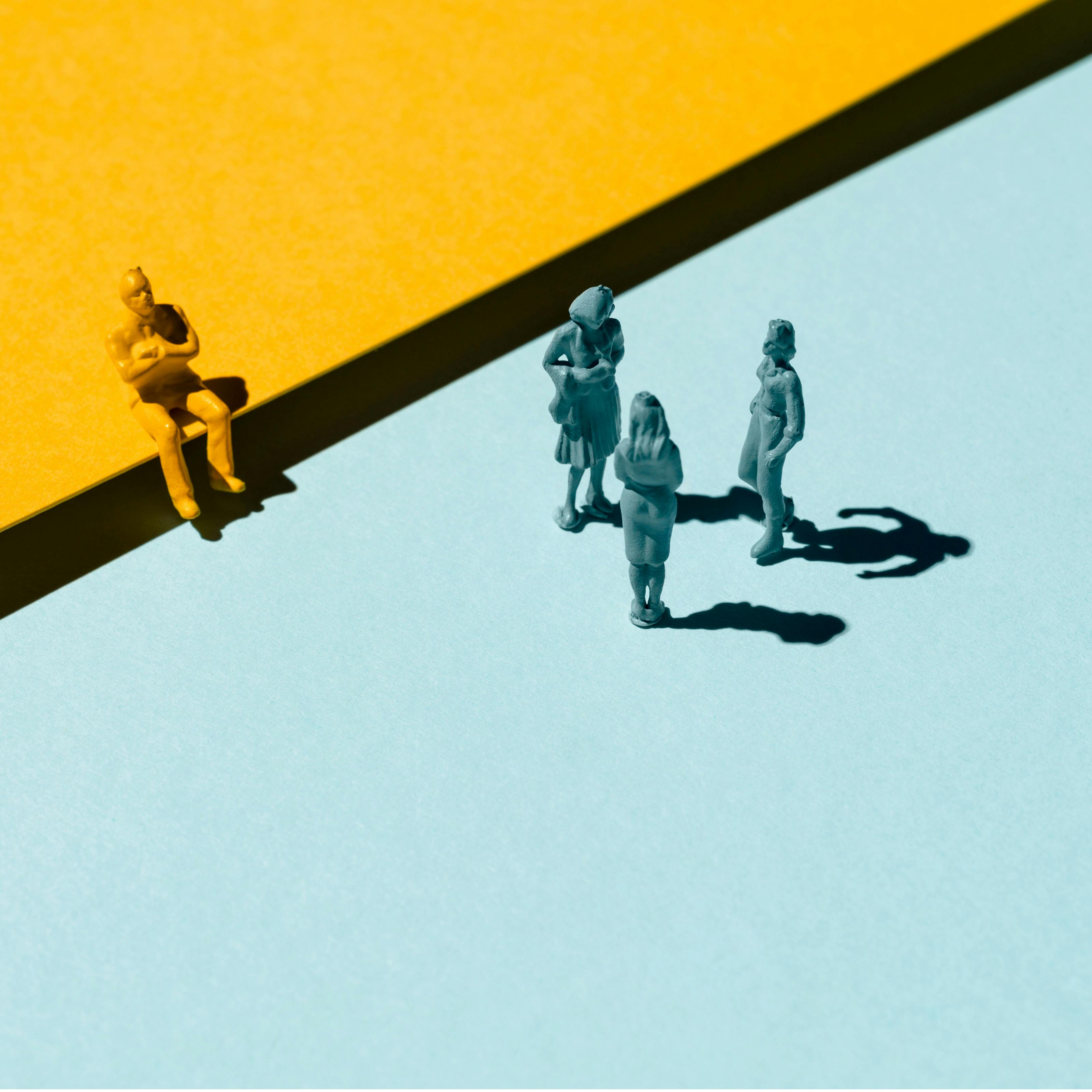 Photograph of a flat surface split between the colours blue and yellow on the diagonal. The yellow surface is raised slightly higher than the blue. A yellow male model figure sits, arms crossed on the edge of the yellow background. Three blue female model figures stand in discussion on the blue background.