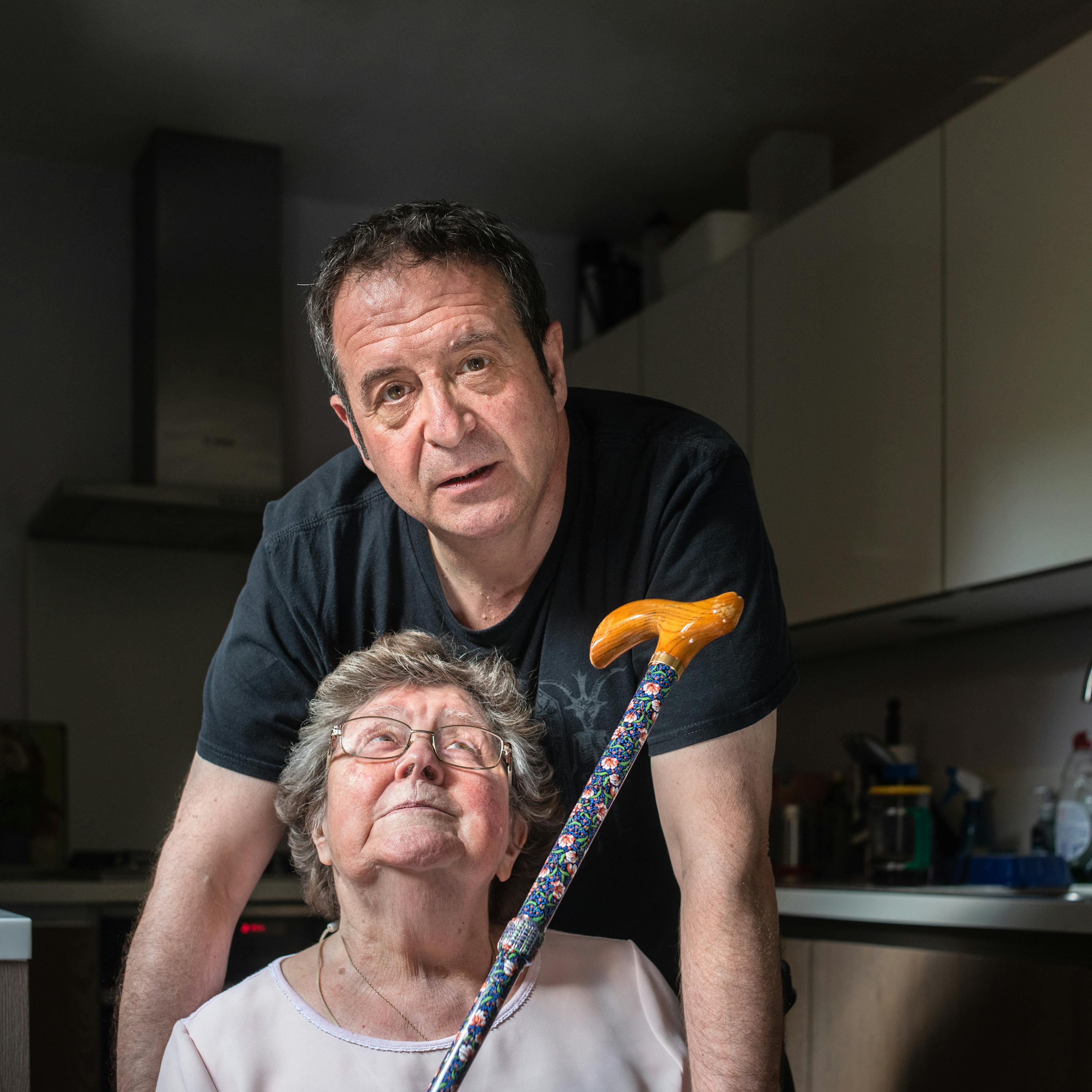 Photograph of a man and his mother in a kitchen. The man wearing a black t-shirt is stood behind his mother who is sitting on a chair. The man is looking straight to camera, leaning slightly forward, his arms resting on the arms of the chair which are out of shot. His mother is wearing a pink blouse and is looking vertically upwards towards him. She is holding her walking stick up towards him, handle end first. In the background are kitchen units, a Tupperware box of biscuits and the tap of the sink.