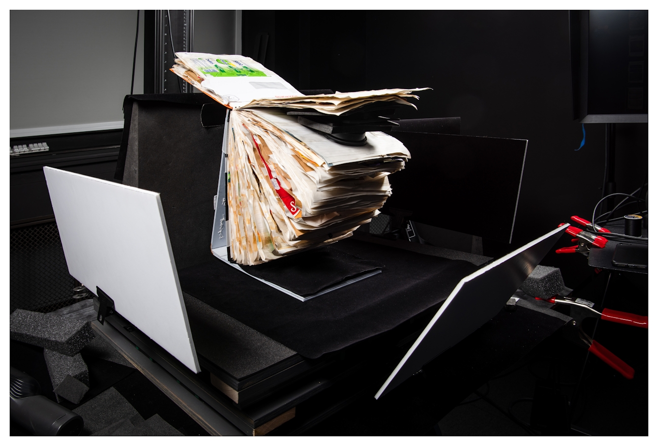 Photograph of a photographic studio flat copy set up, with a large scrapbook on a specialised support, pages open ready to be photographed. Surrounding the scrapbook are black 'flags' and a black background. Around the edge a computer display and red clips are just visible.