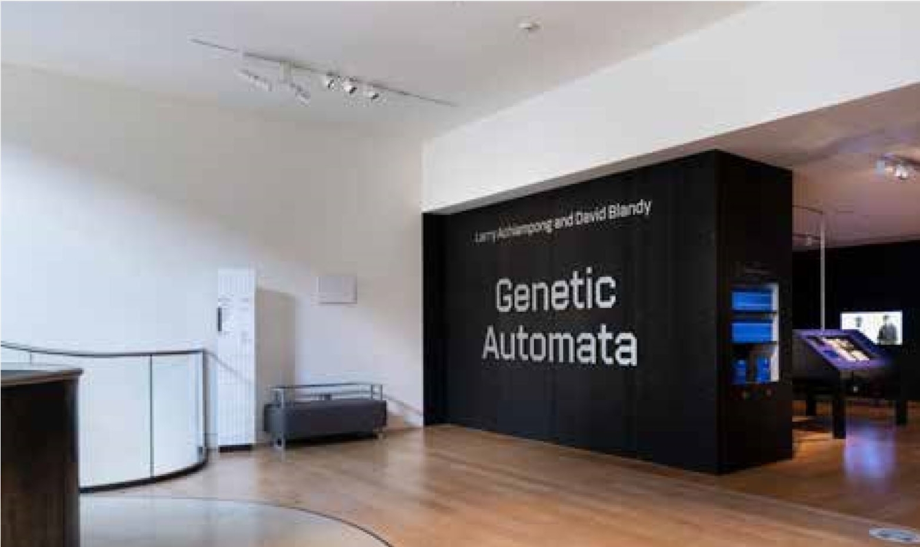 The entrance to an exhibition called Genetic Automata in a modern white-walled interior space with wooden floors and curved glass balustrade.