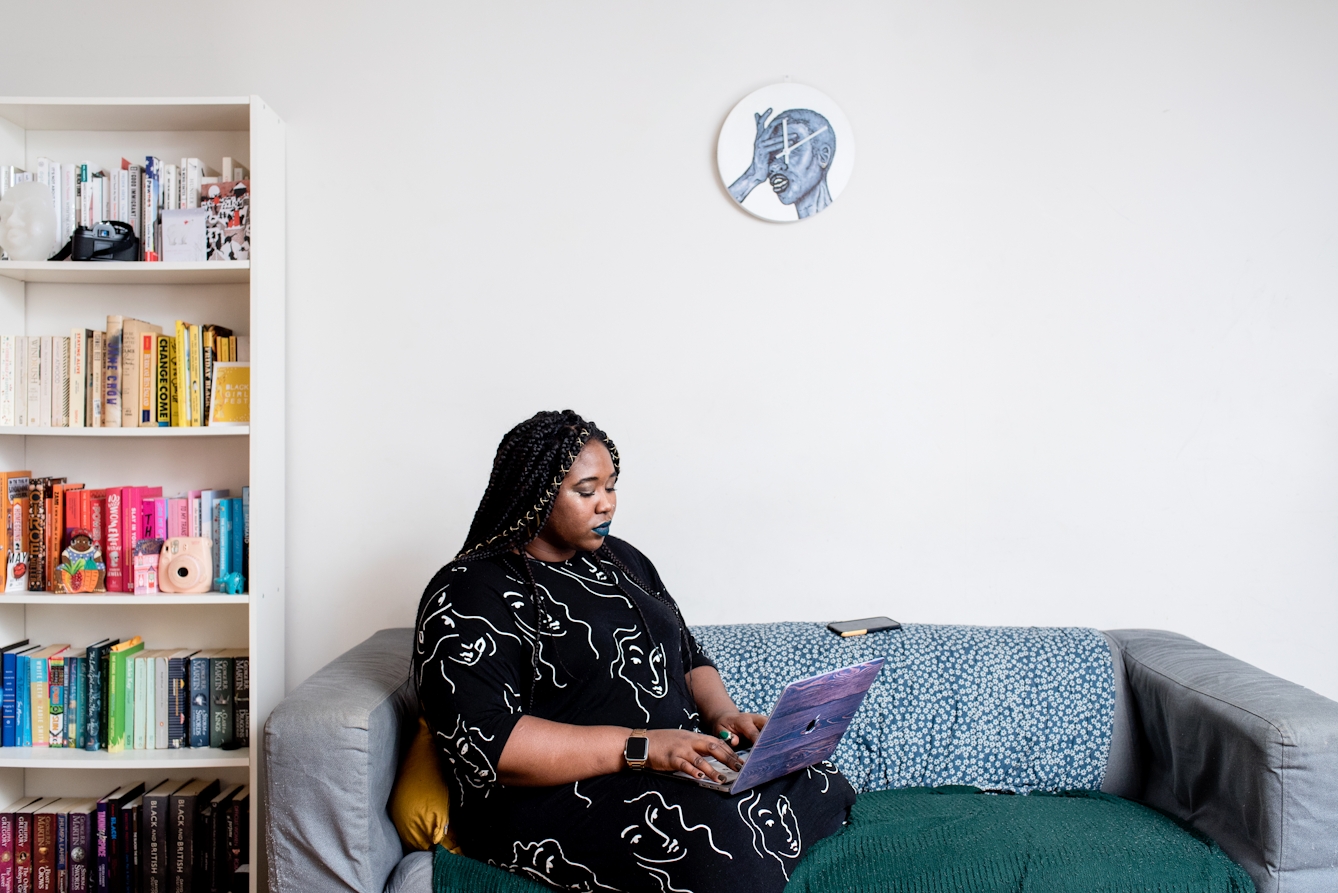 Photograph of a woman inside a house, sitting on a sofa, typing on a laptop computer. To the left is a bookshelf with books on every shelf. Hung on the wall behind her is a clock with an illustration of a face with a hand clasped over the eyes, the mouth slightly open.