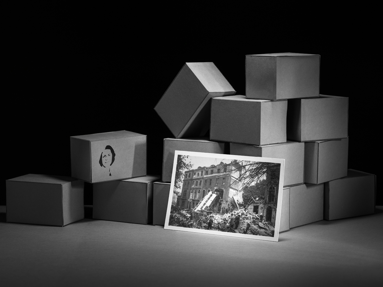Photograph of a set-built scene made up of a grey card horizontal base against a black vertical background. Built into a mock wall are many rectangular brick blocks also made out of grey card. The bricks are stacked together in an untidy pile. Leaning up against the brick wall is a photographic print showing the destruction of London in the Blitz. Stencilled onto the side of one of the boxes is a portrait of a young woman with short hair.