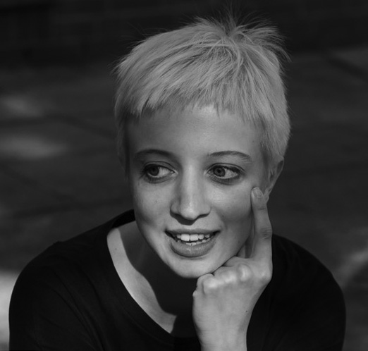 Head and shoulders photo portrait of a young woman with short platinum blond hair, resting her chin on her left hand with her index finger raised and touching her cheek.