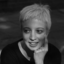 Head and shoulders photo portrait of a young woman with short platinum blond hair, resting her chin on her left hand with her index finger raised and touching her cheek.