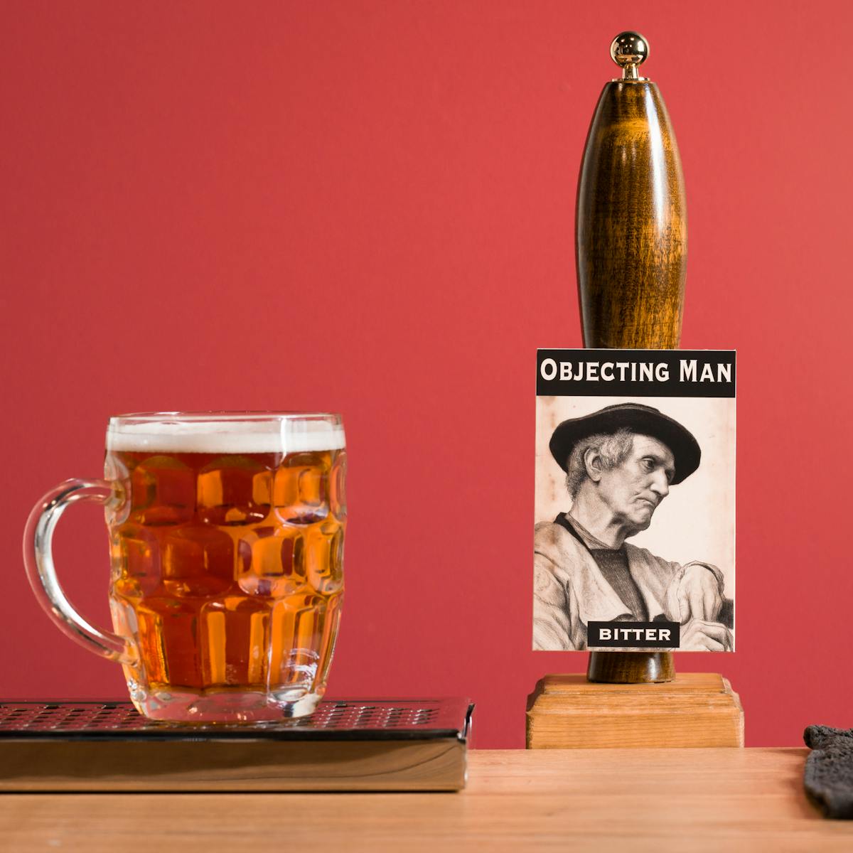 A photograph of a pub bar with an ale pump, drip trays and a full pint of beer in a dimpled glass with a handle. The pump has a badge on it depicting an old white male and the text ‘Objecting man Bitter’.