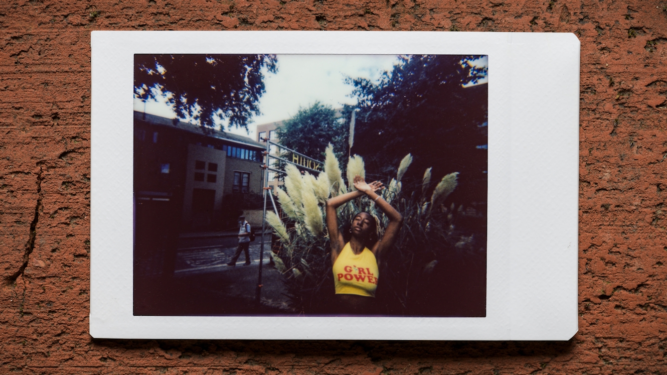 Photograph of an Instax Mini instant film print resting on a textured brick surface.  The print shows a woman standing against the branches and leaves of trees and shrubs outside a street scene and a row of buildings. She has her arms raised up above her head to show her armpit hair. She is wearing a yellow crop top with the words 'Girl Power' written in red across her chest. In the background someone can be seen walking past.