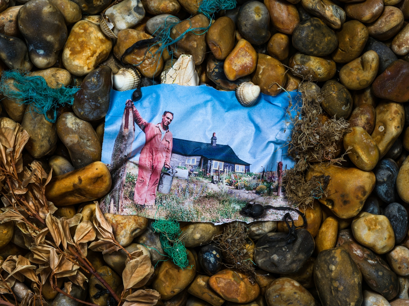 Photograph of a shingle beach made up of pebbles of all different sizes and shapes with a mix of brown and yellow hues. Nestled in the pebbles is a colour photographic print, soaked in sea water such that it has taken on the shape of the stones beneath. The print show Derek Jarman standing in the garden of his Dungeness cottage. With one hand he is holding on to a vertical piece of driftwood, the other holds a galvanised watering can. He is bathed in sunlight, smiling to camera. Surrounding the print on the stones are sea shells, sprigs of beachfront vegetation and the frayed blue and green nylon threads of fishing nets.