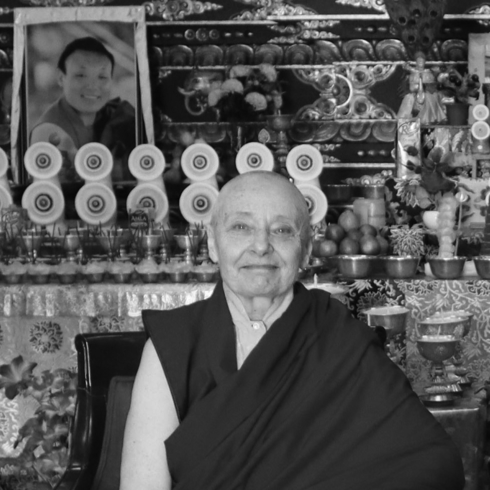 Black and white, head and shoulder photograph of a bald woman wearing a dark robe. In the background there are various ornaments and a photo frame.