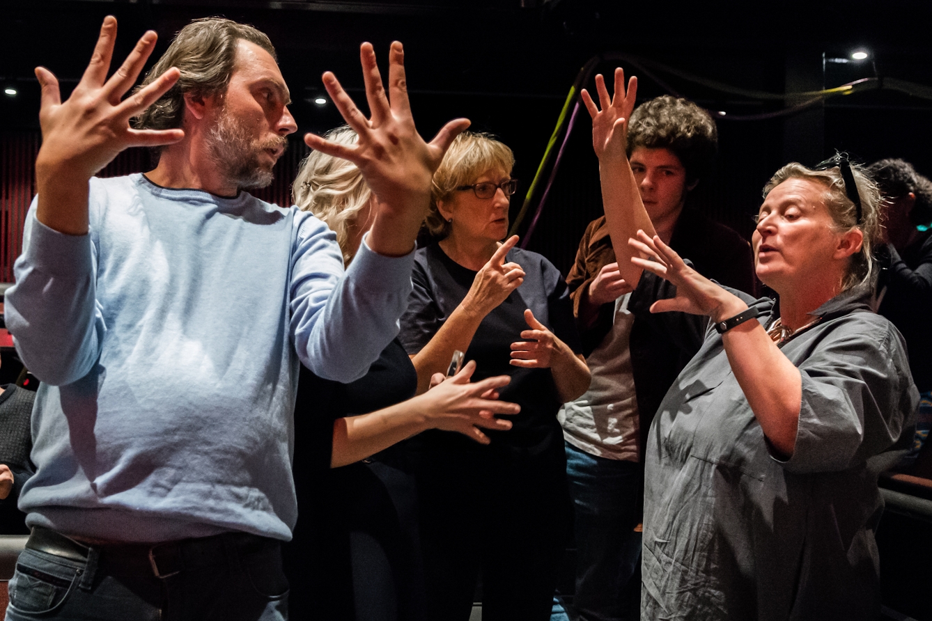 Photograph of a group of people rehearsing. In the foreground to the left of the frame a man with his body facing the camera, his arms out before him and palms facing inwards, his fingers outstretched in a dramatic fashion.  He is looking over his left shoulder towards a woman who has her arms out before her, as if explaining what his movements should be.  Behind them is another group of people who are listening to the instruction.
