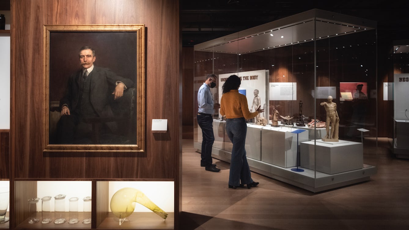 Photograph of two people standing in a gallery looking at and discussing material on display in a glass case, in the foreground of the image is an oil painting of Henry Wellcome.