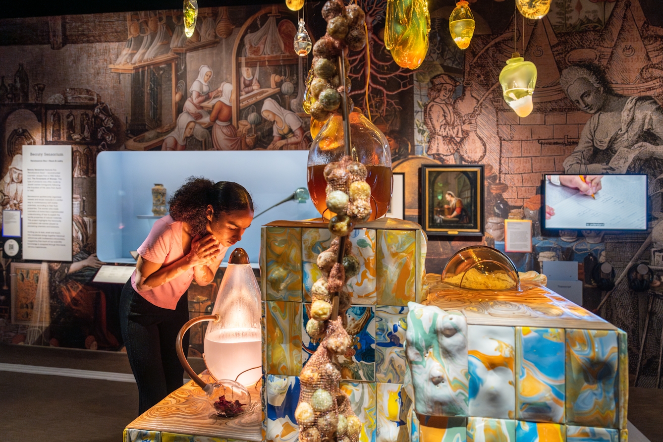 Photograph of a gallery visitor leaning over and smelling a large glass vessel within an installation. At the centre is multi-levelled podium covered in ornate coloured tiles. Suspended above are organically shaped glass vessels containing coloured liquid and natural substances. The wall behind in covered in renaissance images. The whole scene is dark but warmly lit.