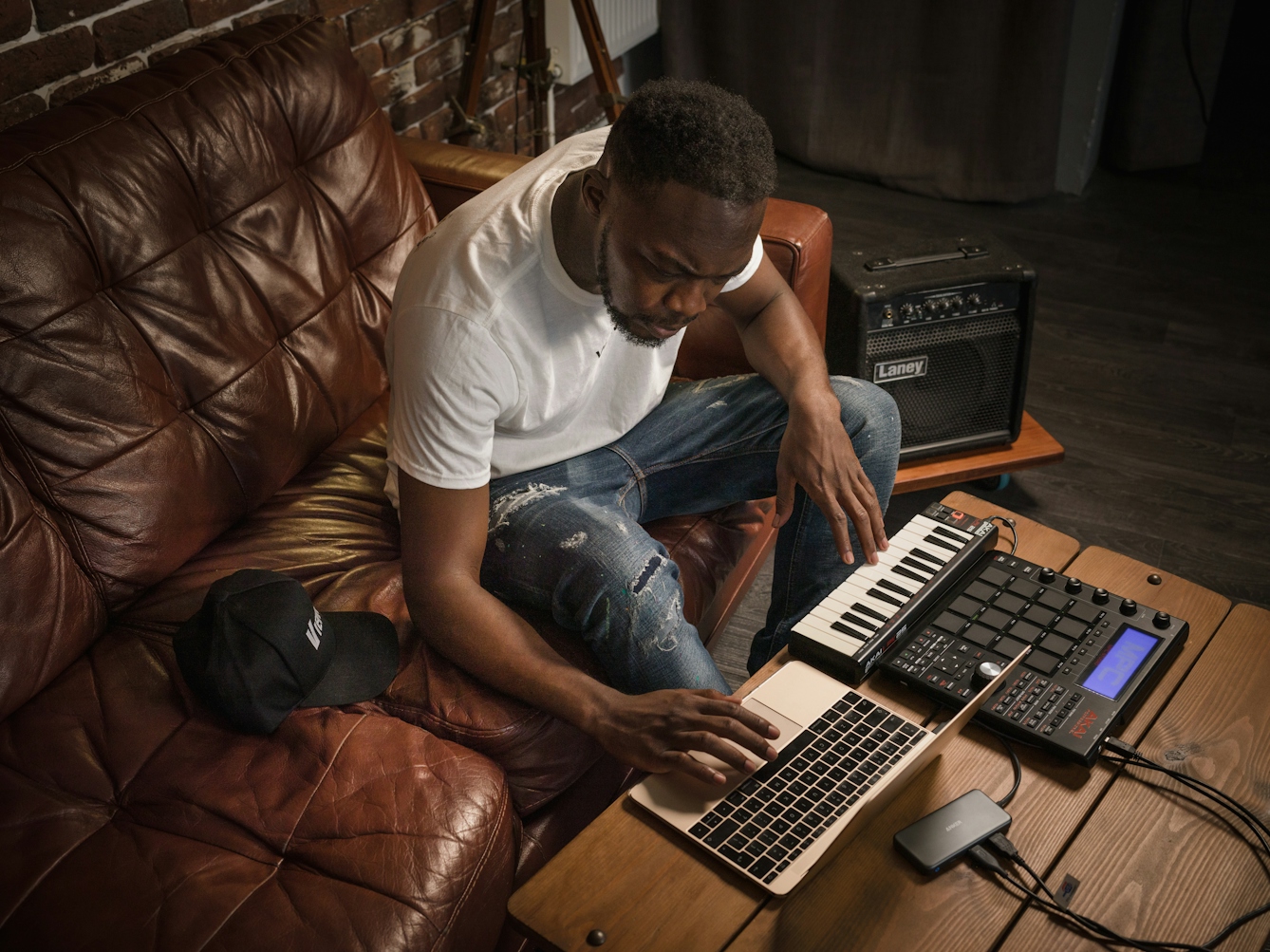 Photograph of a man in a white t-shirt sitting on a brown leather sofa whilst simultaneously playing a note on his keyboard and using his laptop. On the table in front of him is a keyboard, laptop and beatbox, whilst a large bass speaker is to his left.  A black hat is situated on the sofa to his right.