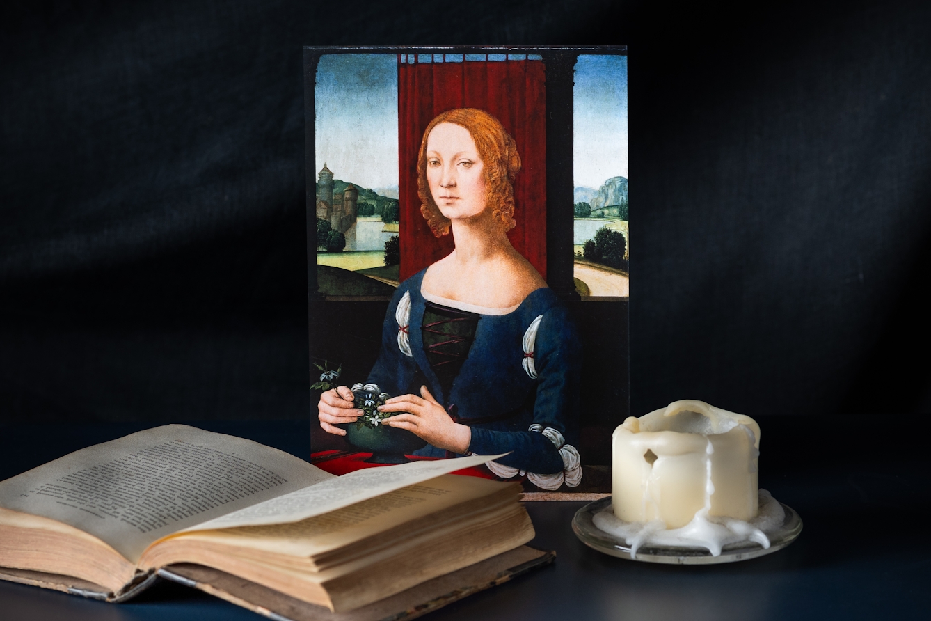 A still-life photograph showing a reproduction of a painting depicting a Renaissance women. She has curly red hair and is dressed in blue with white details. There is a red curtain behind her with a view of countryside in the background. She is tending to a bowl of small white flowers. Next to the painting is an opened book and a large candle with wax dripping down the sides.