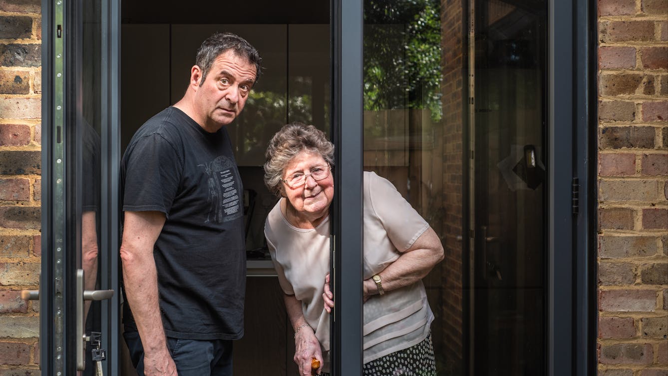 Photograph of a man standing in a glass patio doorway with his mother, both are looking to camera. One of the doors is open. The man is slightly hunched forwards, eyebrows raised causing his brow to furrow. He looks a little 'long suffering'. His mother is peeping around the large glass patio doors, holding a walking stick in her right hand and the door frame in her left. She has a cheeky smile on her face. Either side of the patio doors is the brickwork of the back of the house.
