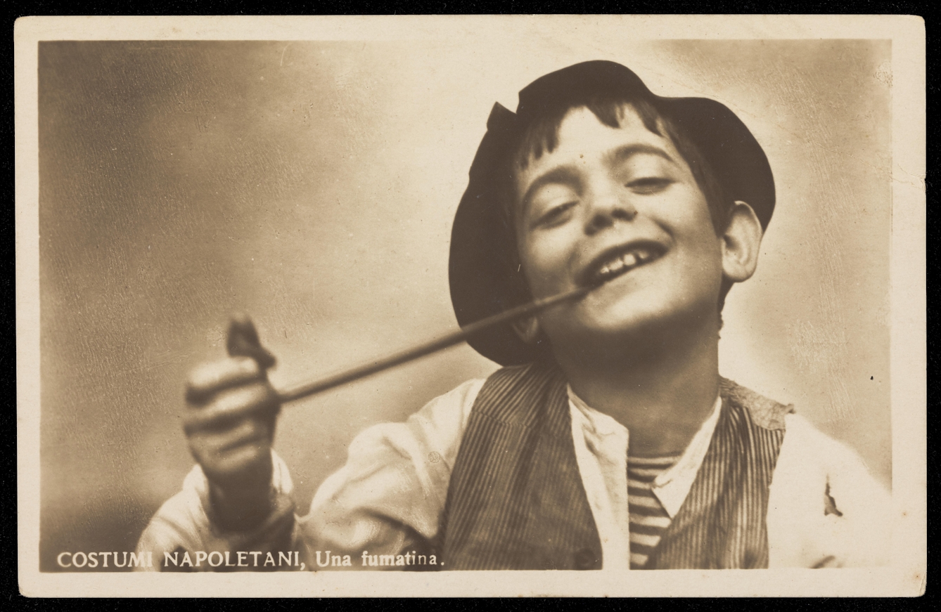 A black and white photograph with sepia hue. It shows the head and shoulders of a boy. The boy is wearing a dark hat, striped waistcoat and white shirt. He smiles while holding a long pipe in his mouth.
