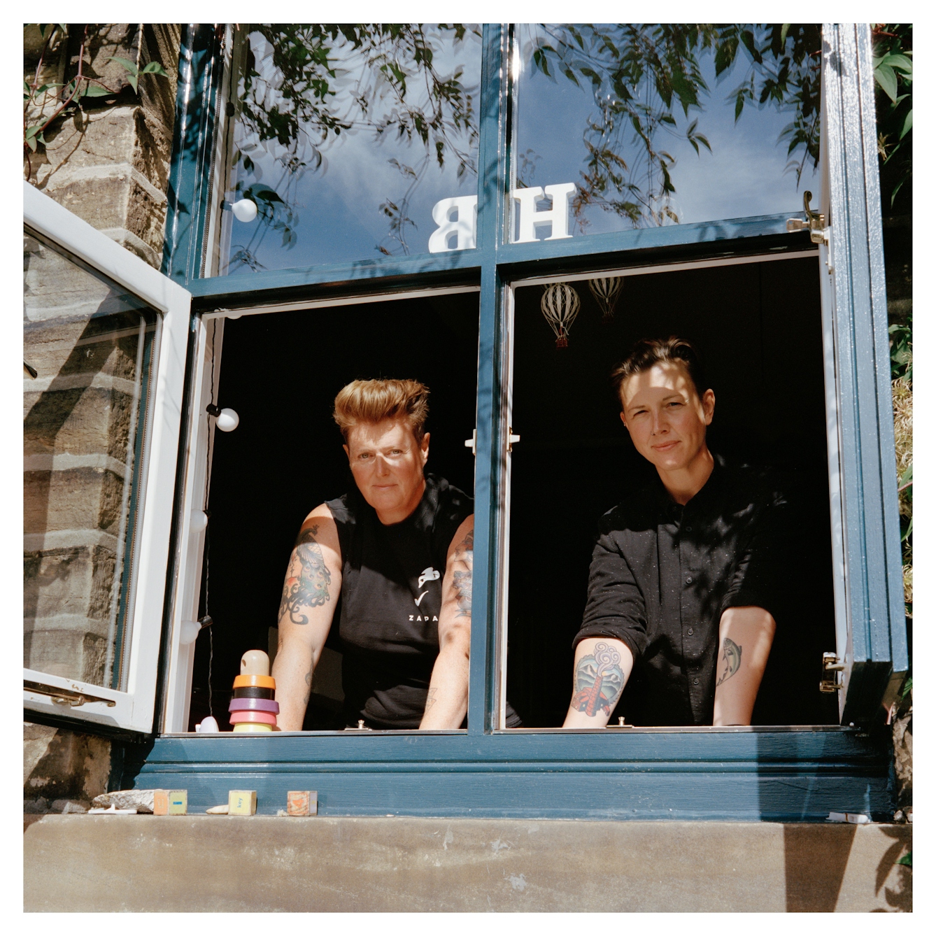 Portrait photograph of Belinda and Heidi standing in an open window, looking outside towards the camera. The woman on the left has short brown hair and is wearing a black sleeveless top, she has several tattoos on her arms. The woman on the left has short dark hair and is wearing a black shirt - she has several tattoos on her arms. 

They are both looking directly at the camera. There is a stack of colourful wooden rings on the windowsill and above their heads, a wooden initial H and B. 
