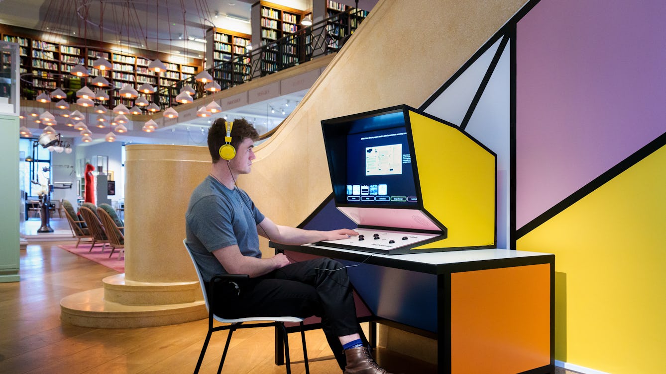Photograph of a gallery installation showing a desk set in the corner of a larger gallery space. On the desk is a large console made up of a screen below which are a series of black arcade style buttons. The console has a bright yellow side and bright pink front. Seated at the desk is a young man wearing jeans and a t-shirt. He is watching a film playing on screen and is resting his left hand on the button panel. On the screen are various rectangular graphic elements containing text. The walls behind the desk are white with strong graphic black lines crossing them in a geometric pattern. One of the spaces created by the intersecting lines is coloured pink, another yellow. In the distance to the left, the rest of the gallery can be seen, made up of book shelves, display cases and a series of hanging lampshades.