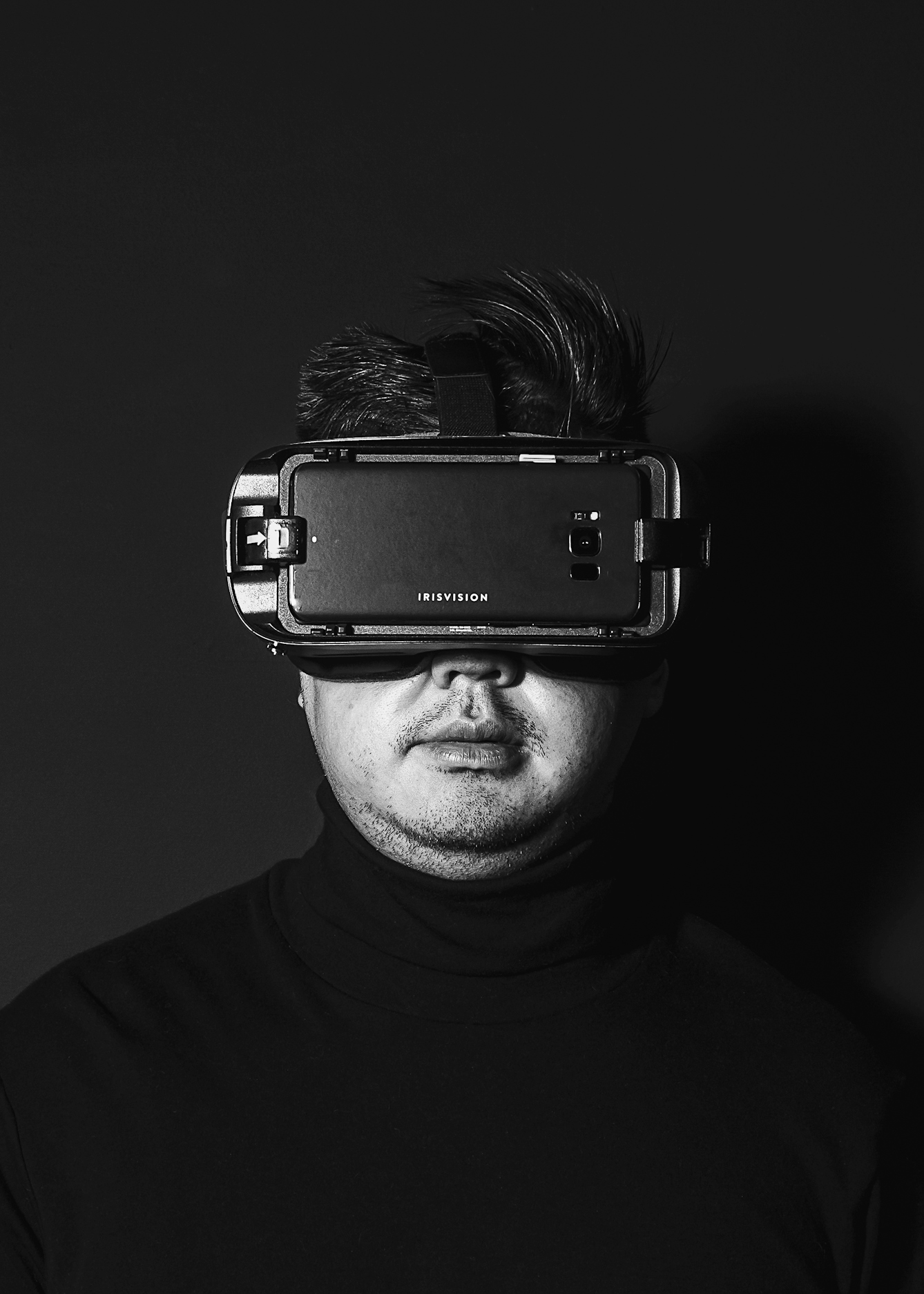 Black and white photographic portrait of a man from the chest up looking to camera. He is wearing a set of virtual reality goggles and a polo neck black jumper. He is spotlit in a small circle of light. He is standing against a black background which means he is surrounded by darkness.