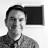 Black and white photograph of a white man looking directly at the camera and smiling. A framed black square is behind him.