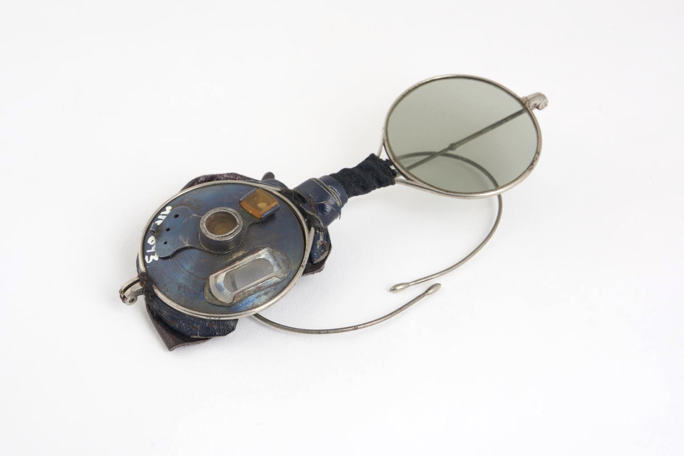 Circular, wire-framed spectacles with coiled arms to clip around the ears. The right single glass lens has been replaced iwth a metal disc holding three small lenses, one of orange glass. The bridge of the spectacles is wrapped in a compination of leather and cloth.