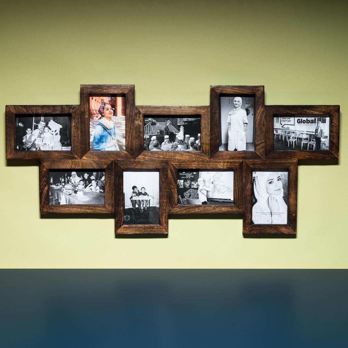 Photograph of a multi-frame photo frame containing nine photographs, eight in colour and one in black and white. The frame is hung on a light green coloured wall above a blue tabletop.