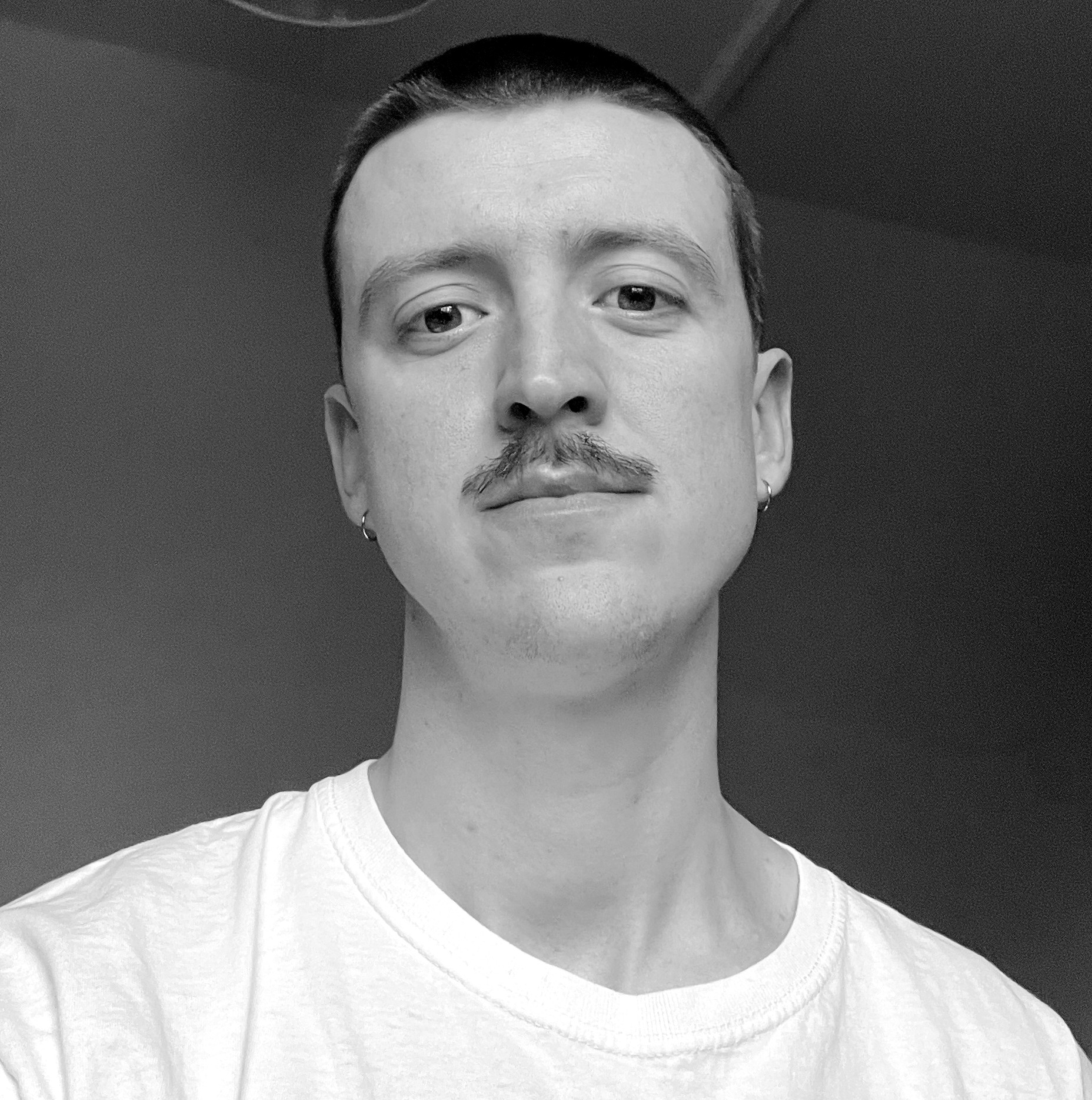 Black and white photograph of the head and shoulders of a young white man with a mustache and short cropped hair. The man is looking straight into the camera