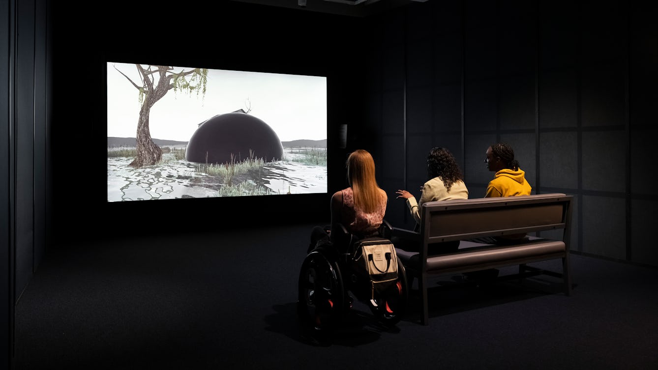 Photograph of a dark exhibition space featuring a projected video on a large screen. Three visitors are watching the film. 