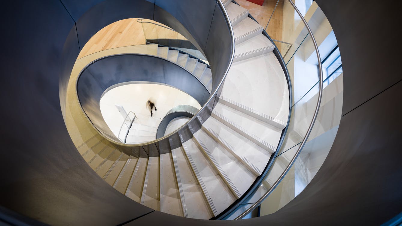 Photograph looking from up high down the centre of an irregular spiral staircase. The sides of the staircase are grey metal with cream stone steps. In the distance on the ground floor of the staircase is a small dark blurred figure about to start climbing the stairs.