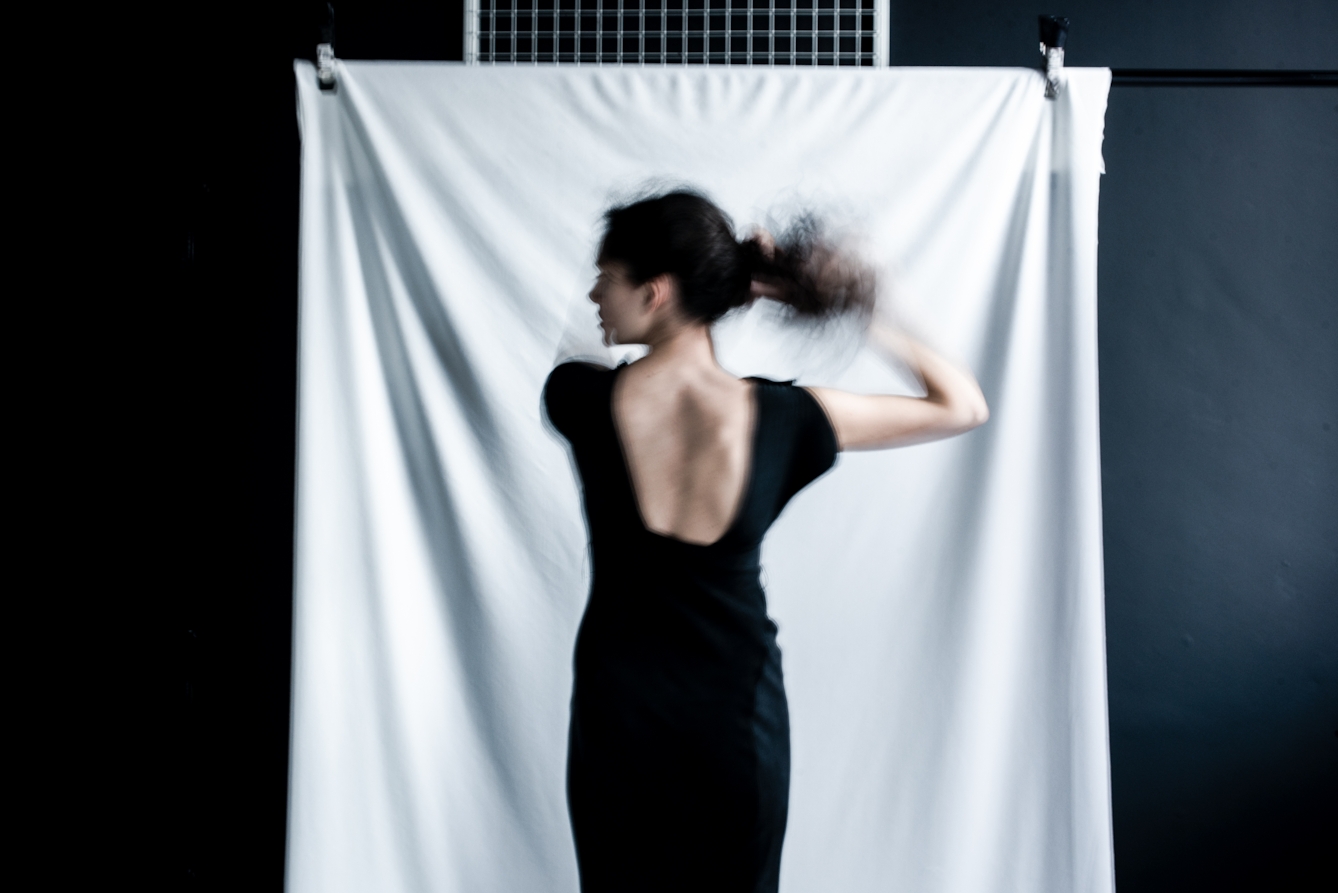 Photograph of a young woman from behind, from the thigh up. Her head is turned to the left revealing her profile. She is wearing a low cut black dress and is in the process of tying her hair. The long exposure of the camera has captured her movement. In the background is a drape of white fabric.