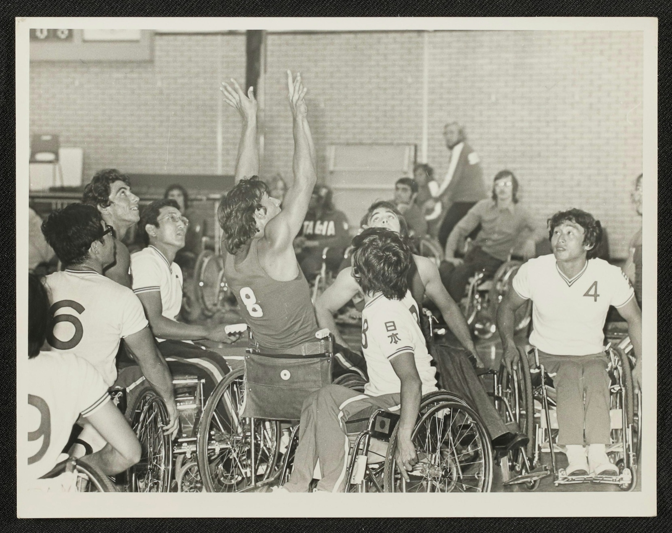 Black and white photographic print showing a wheelchair basketball match. Lots of players are clustered close together in the photograph, looking up into the air. In the centre, a man wearing a dark number 8 shirt has his arms flung upwards as though he has just released the ball for a shot at the net. In the background, spectators seated in front of the brickwork walls look towards the action.