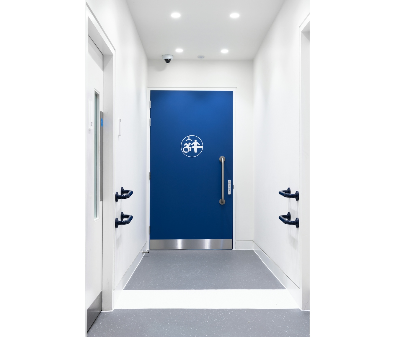 A photograph of the Changing Places toilet with a white logo showing a wheelchair, adult changing table, and a hoist on a large blue door with parallel blue hand rails on adjacent walls.