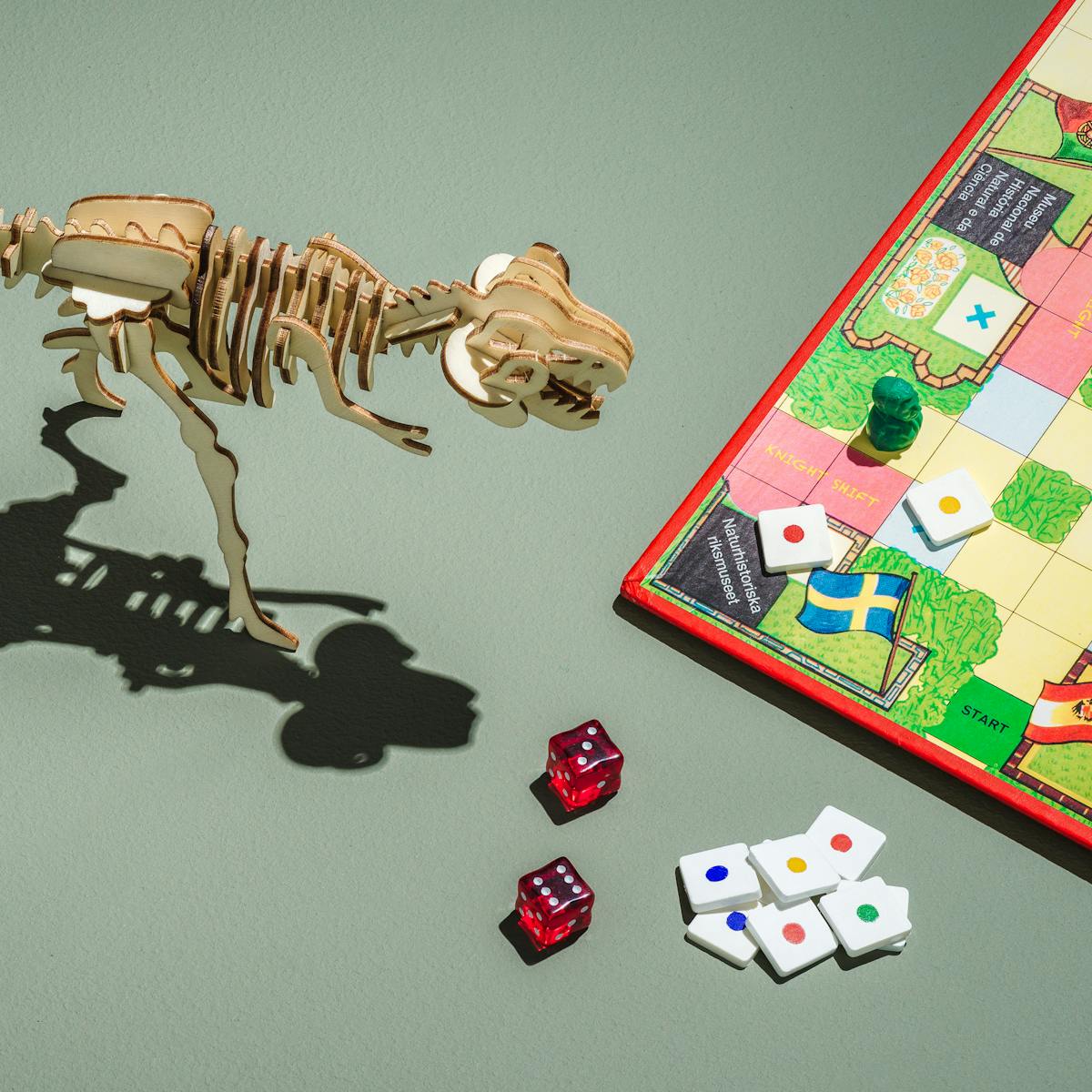 Photograph of a board game placed to the left of the frame,  featuring museums from various countries, red, green, blue and yellow counters, and two red dice.  In the centre of the frame is a laser cut wooden tyrannosaurus rex model.