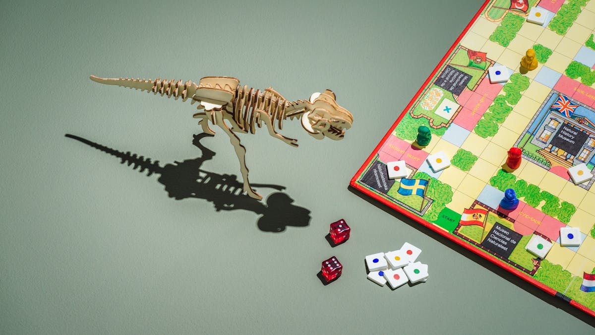 Photograph of a board game placed to the left of the frame,  featuring museums from various countries, red, green, blue and yellow counters, and two red dice.  In the centre of the frame is a laser cut wooden tyrannosaurus rex model.
