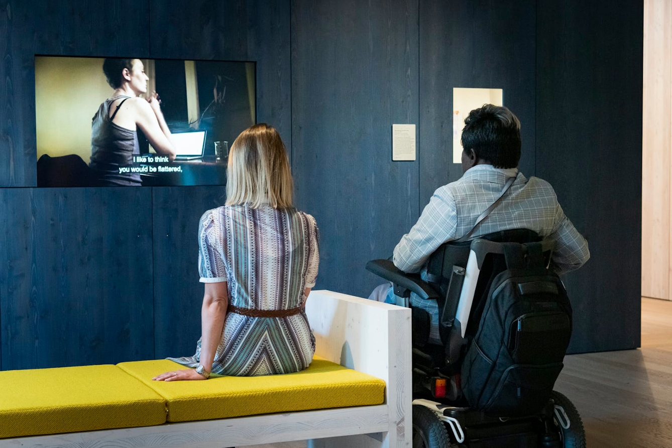 Image showing two visitors watching a film on a screen in the Being Human gallery. One visitor sits on the bench and one sits in a wheelchair alongside. The bench seat is positioned offset to the centre of the screen so that both viewers have a centred viewpoint.