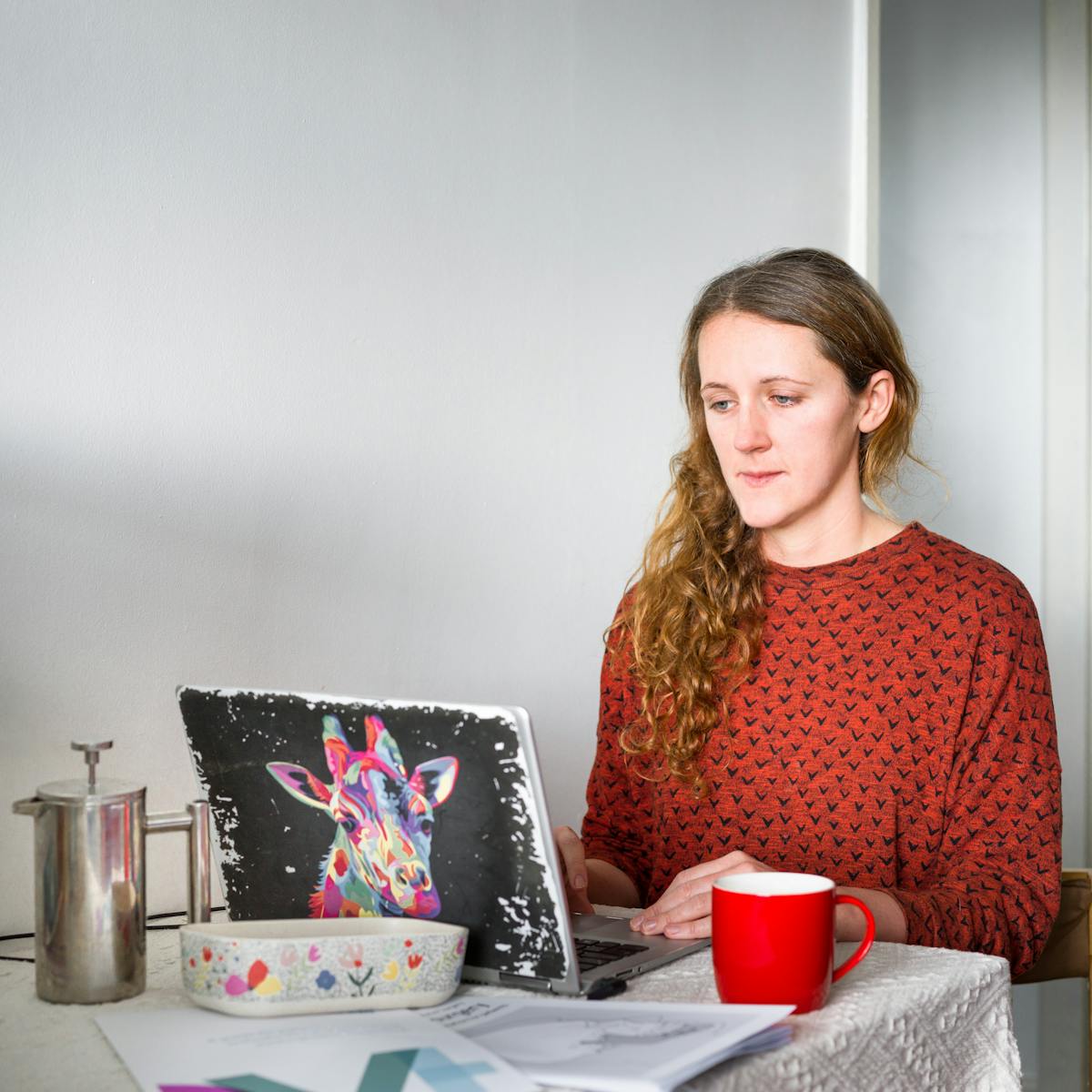 Photographic portrait of a woman sitting at a dinning table against a white wall, in a domestic setting. She is looking down at a laptop screen which is open on the table. Her hands are resting on the keys, in the process of typing. She is wearing a rust coloured top with small black 'v' shapes covering it, which look like birds in flight. Her wavy brown hair is swept over her right shoulder. On the table in front of her is a white fabric tablecloth on which is a red coffee mug, a silver cafetière, a floral dish and printed A4 material. On the lid of her laptop is a colourful drawing of a giraffe's head.