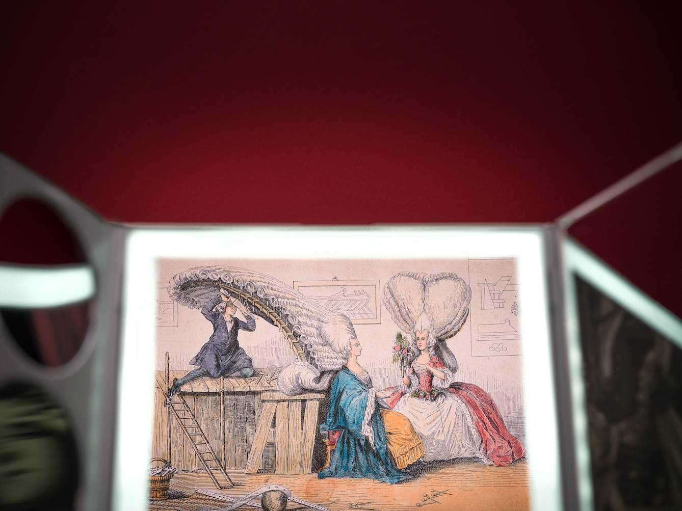 Photograph of the top half of a three-fold beauty mirror against a red background. reflected in the main mirror is a 19th century illustration showing a man tying a woman's absurdly high wig on to a scaffolding; another woman wearing a tall heart-shaped wig looks on.