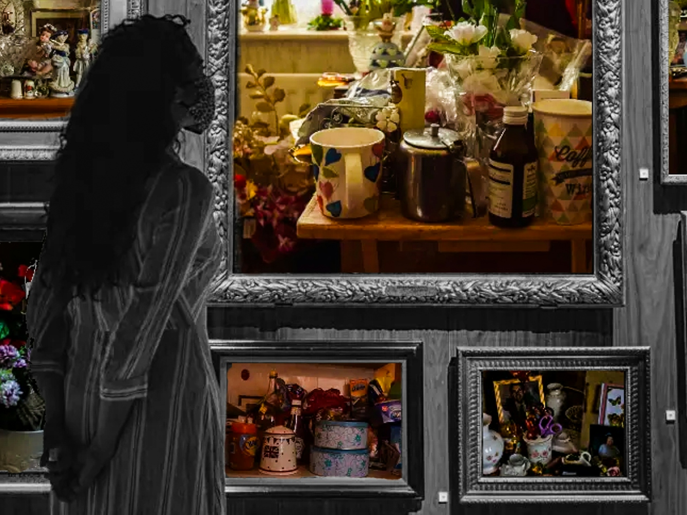 Crop of a black and white photograph showing a wall in a museum covered in large ornate picture frames. In the foreground are two people facing the wall of frames and wearing masks. Inside the black and white frames are colourful photographs showing different household objects. The largest frame in the center shows a table that has lots of things on top of it, all packed very close together, including vases of flowers, a mug and coffeepot, and medicine. On the windowsill behind the table are more flowers. The frame to the right shows a photo of a an equally crowded table, holding photo frames, flowers and various other trinkets. 