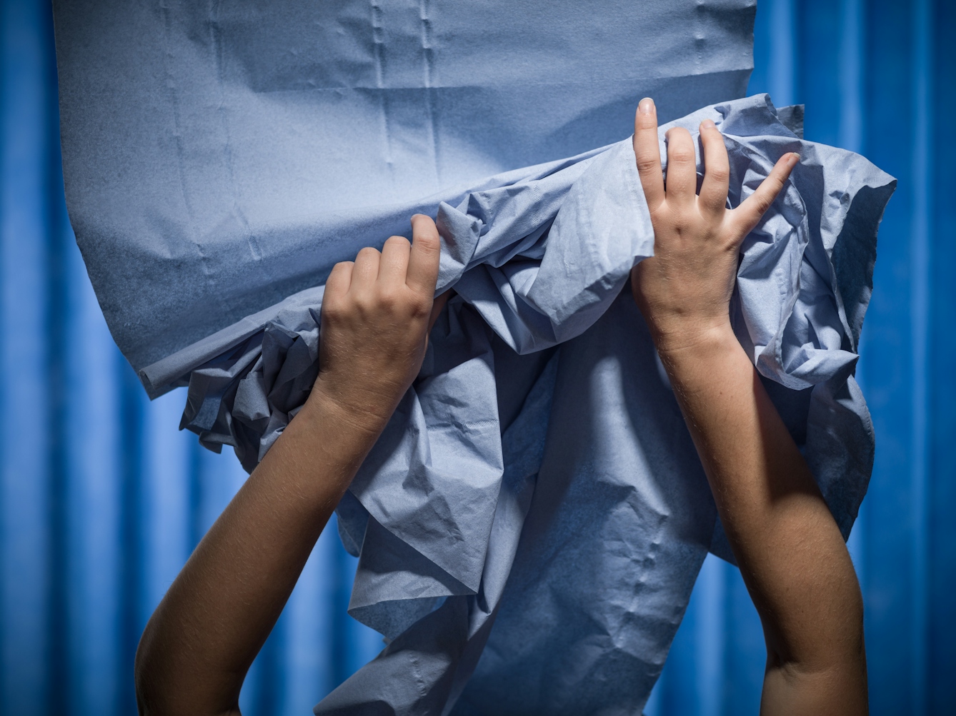 Photograph of a pair of hands struggling to contain a ream of medical blue examination couch paper. Photographed against a background of blue antibacterial clinical curtains.