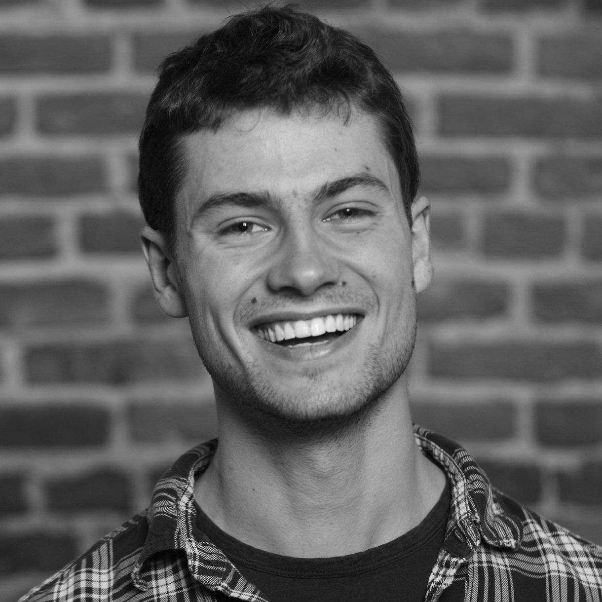 Head and shoulders photograph of a man with shirt hair and even teeth, smiling, wearing a chequered shirt over a black t-shirt. 